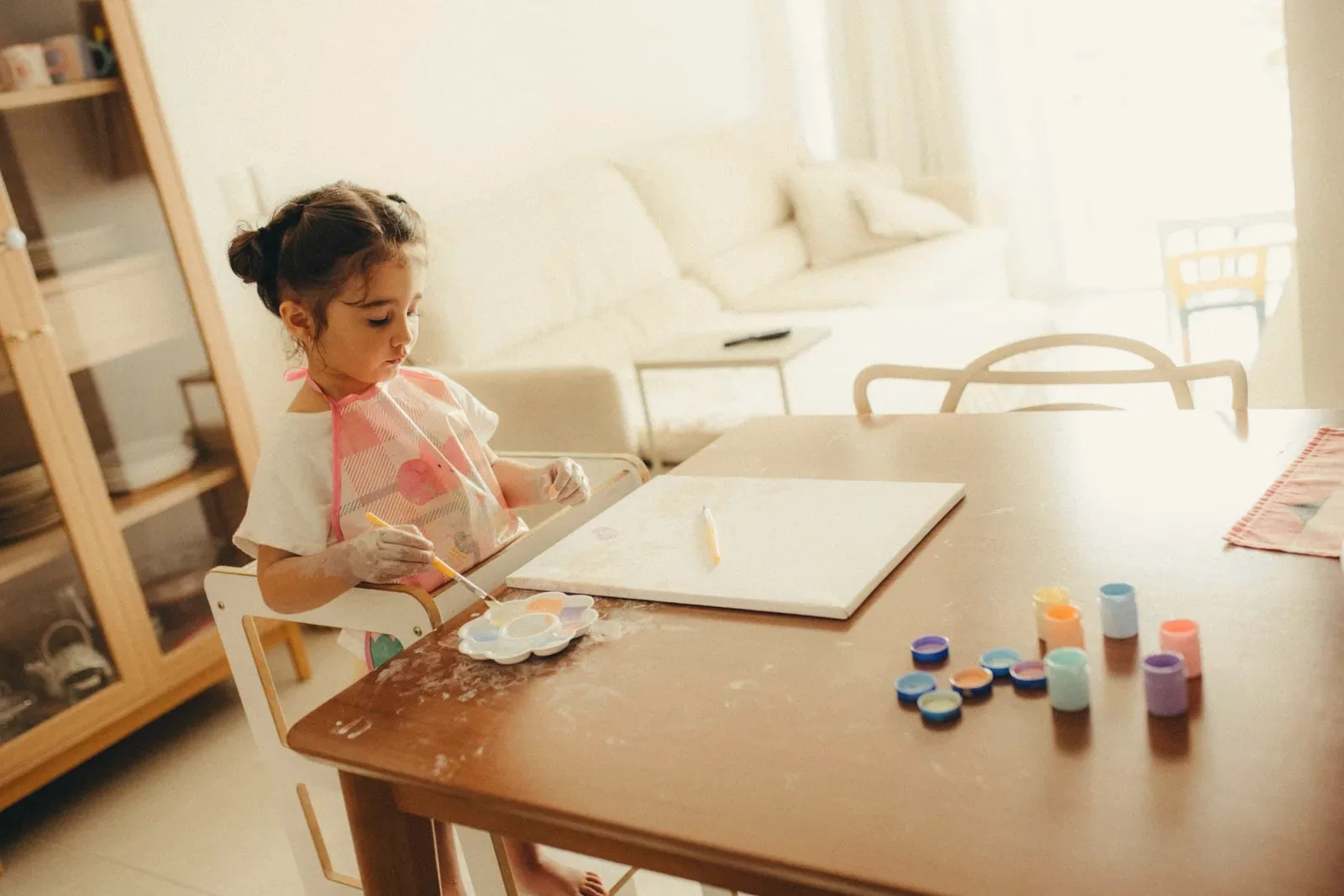 A young girl painting at a dining table in a sun-flooded living room