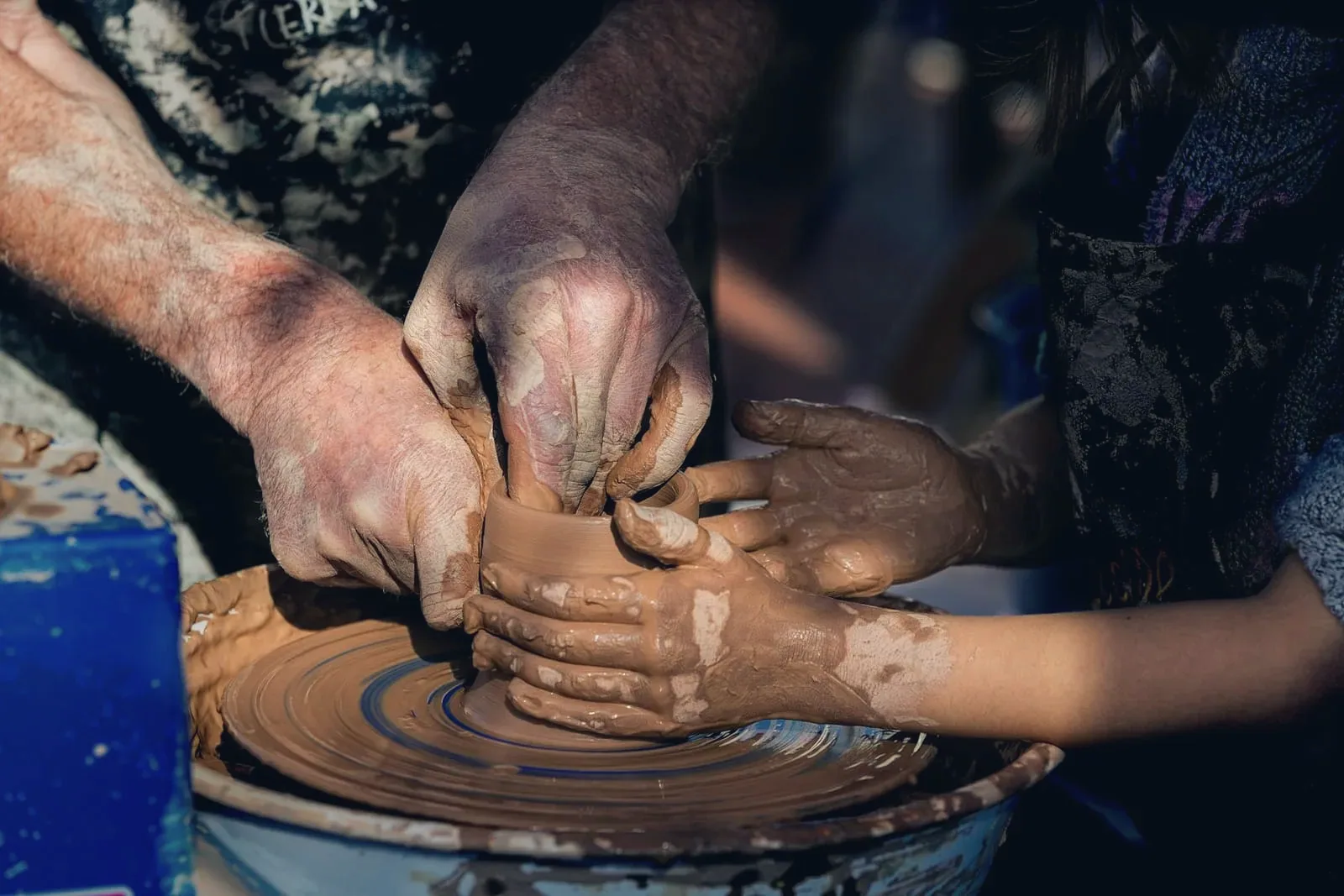 Adult hands guiding a child's clay-covered hands on a pottery wheel