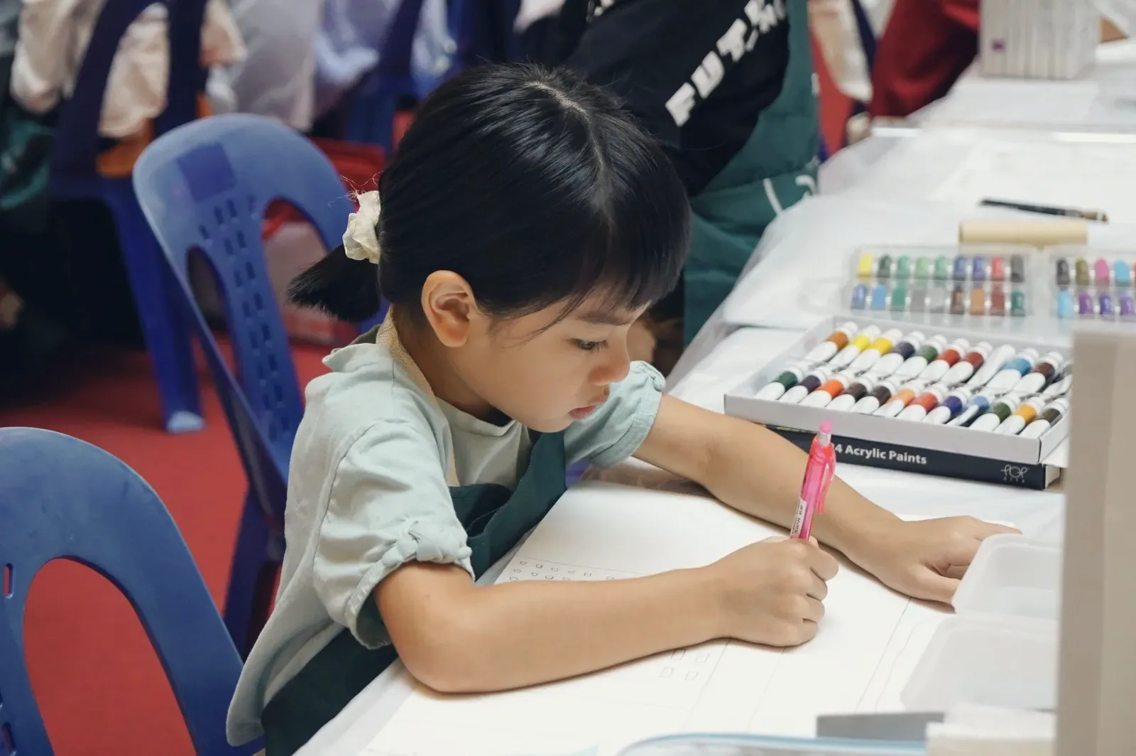 A young student concentrating on her drawing beside a box of acrylic paints