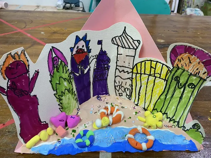 A standing paper cut-out of a seaside town with towers and trees behind a modelled clay beach