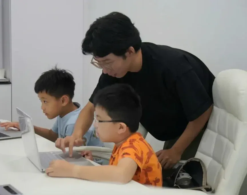 A teacher leans in to guide two boys working on laptops in a coding class