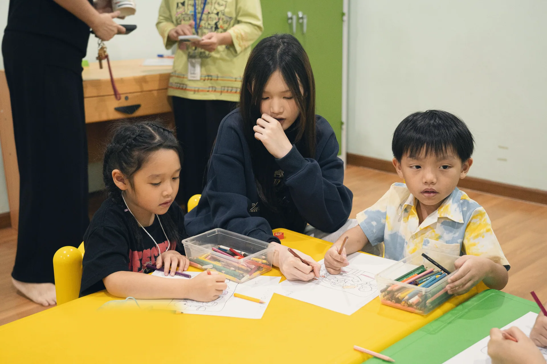 Children drawing with coloured pencils at a yellow table, an older girl sitting with them