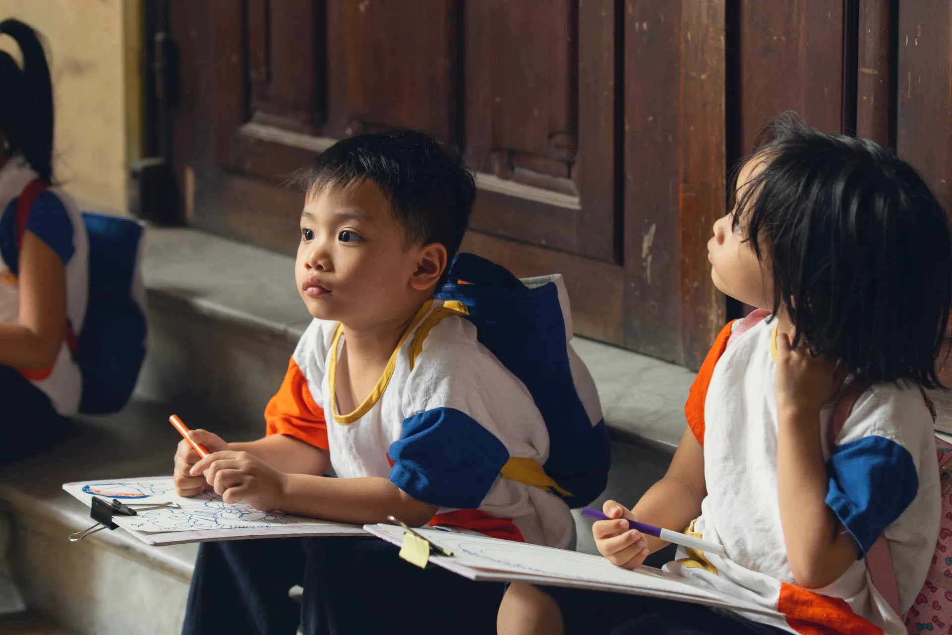 Two young children with backpacks drawing on clipboards