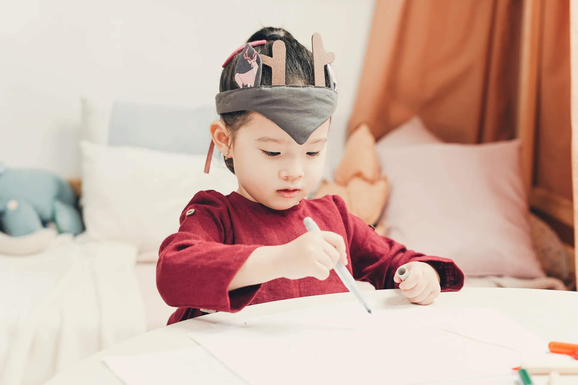 A toddler in a party crown drawing with a marker at a white table