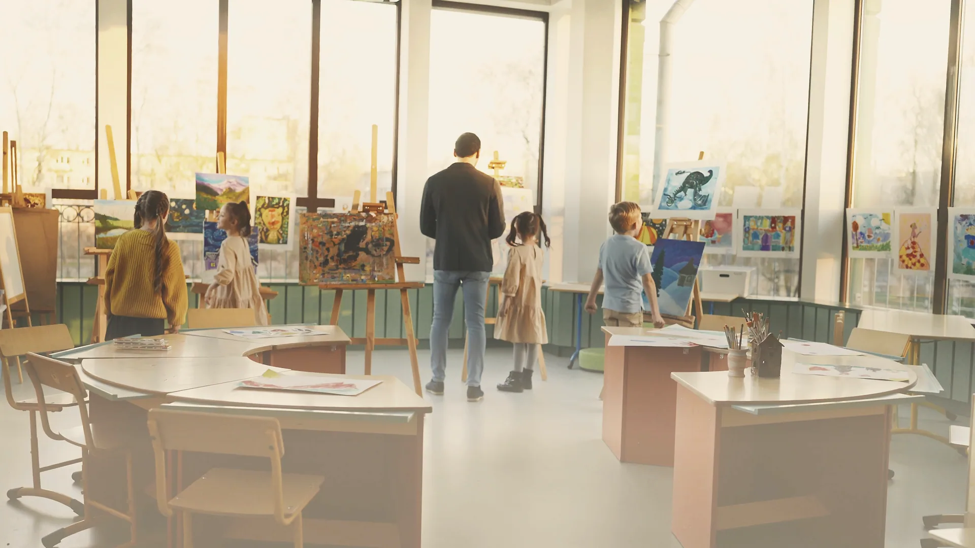 A teacher and four children among the easels in a bright art studio, tall windows of golden light behind them