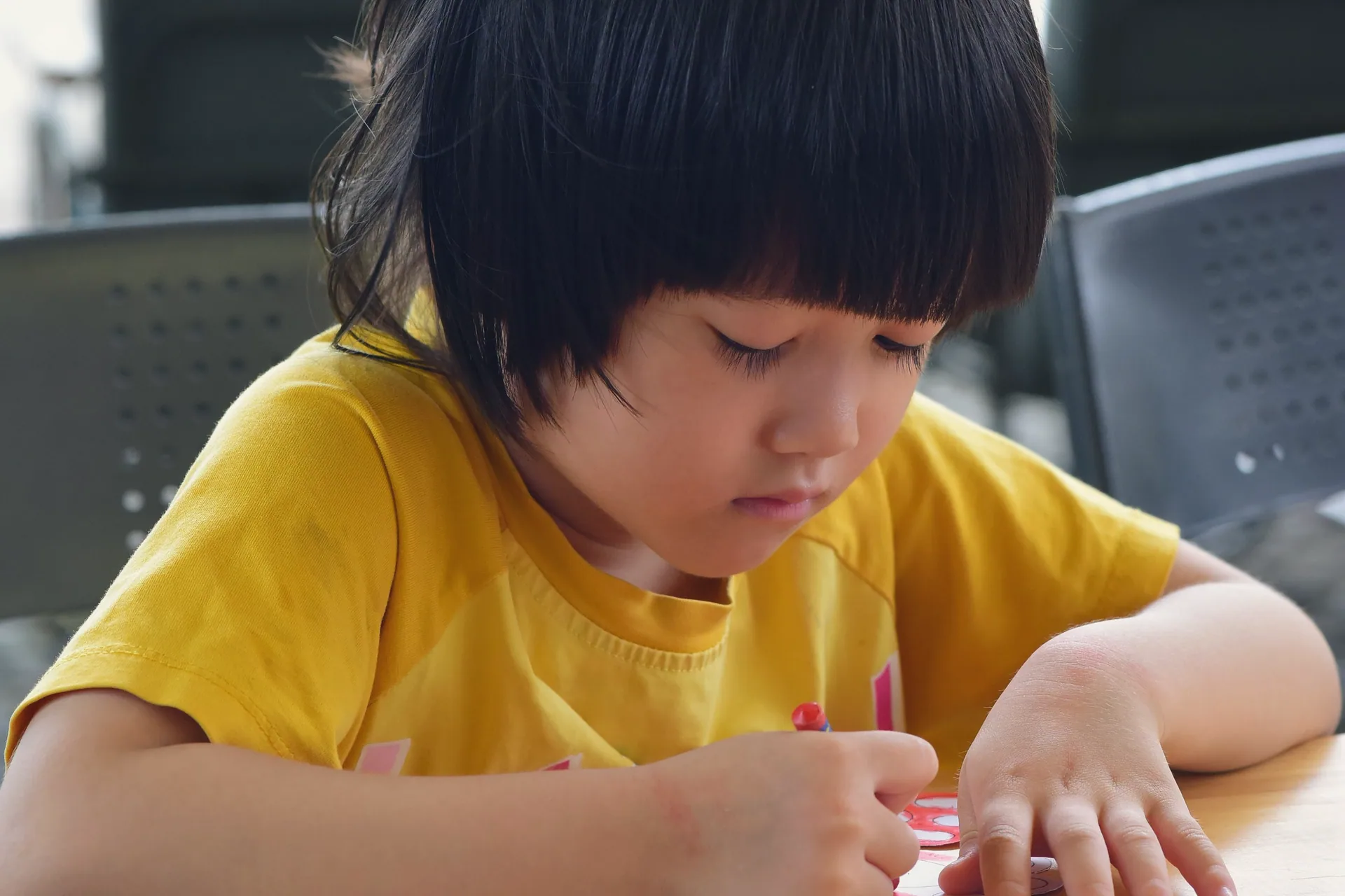 A child in a yellow T-shirt drawing at a wooden table, eyes on the page